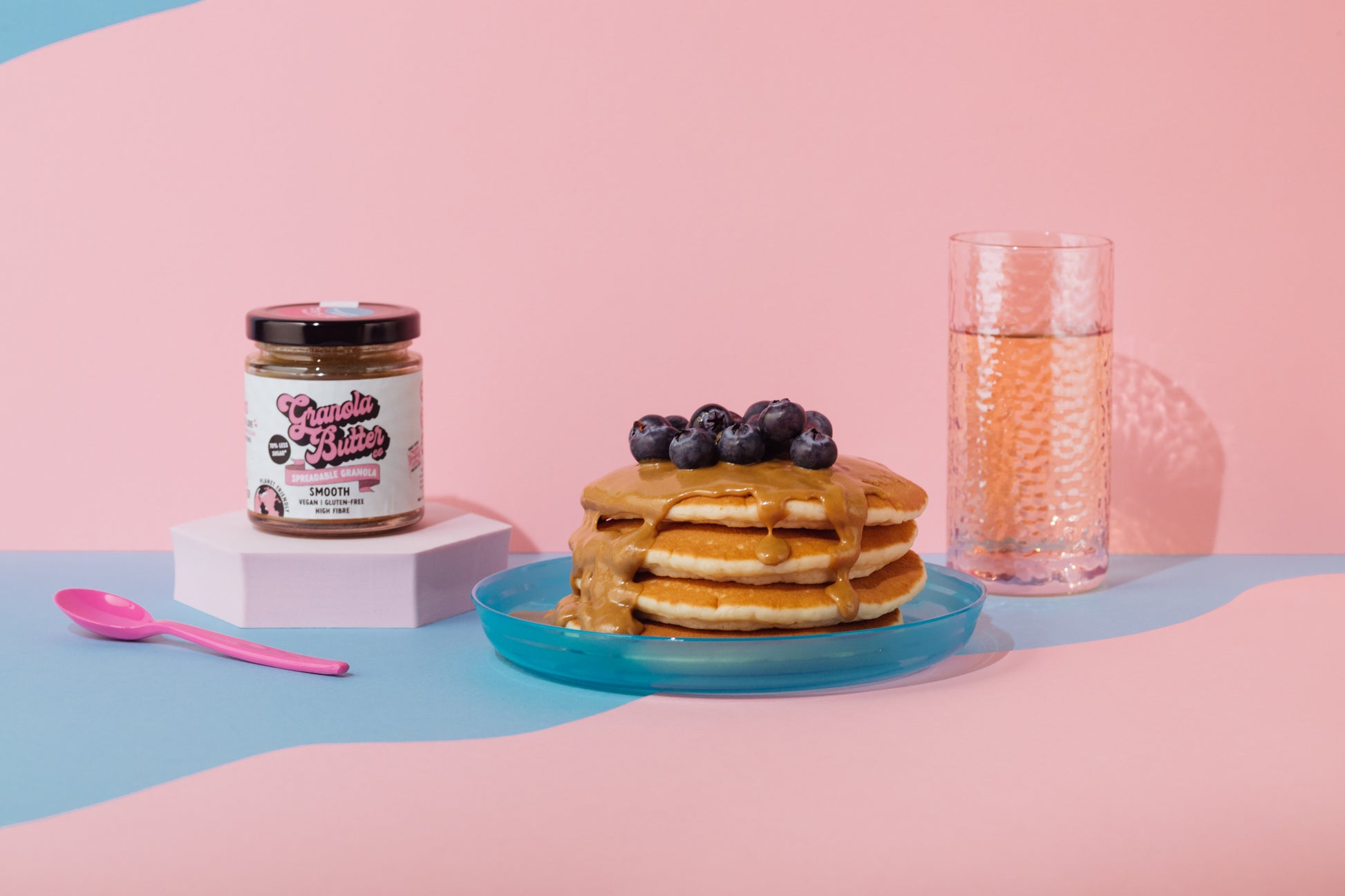 Vegan pancakes stacked and coated in smooth granola butter with blueberries scattered on top. A jar of the sweet spread is placed next to the breakfast delight. 